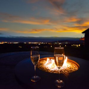Two glasses of champagne placed on a flaming fire pit of a luxury hilltop home's back yard while overlooking a valley and city with lights illuminated at sunset in the evening.